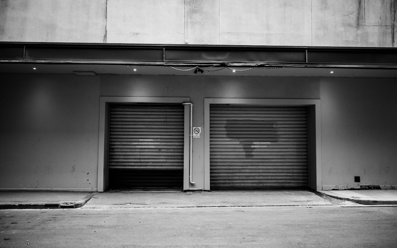 A monochrome urban exterior with two closed garage doors and a concrete wall.